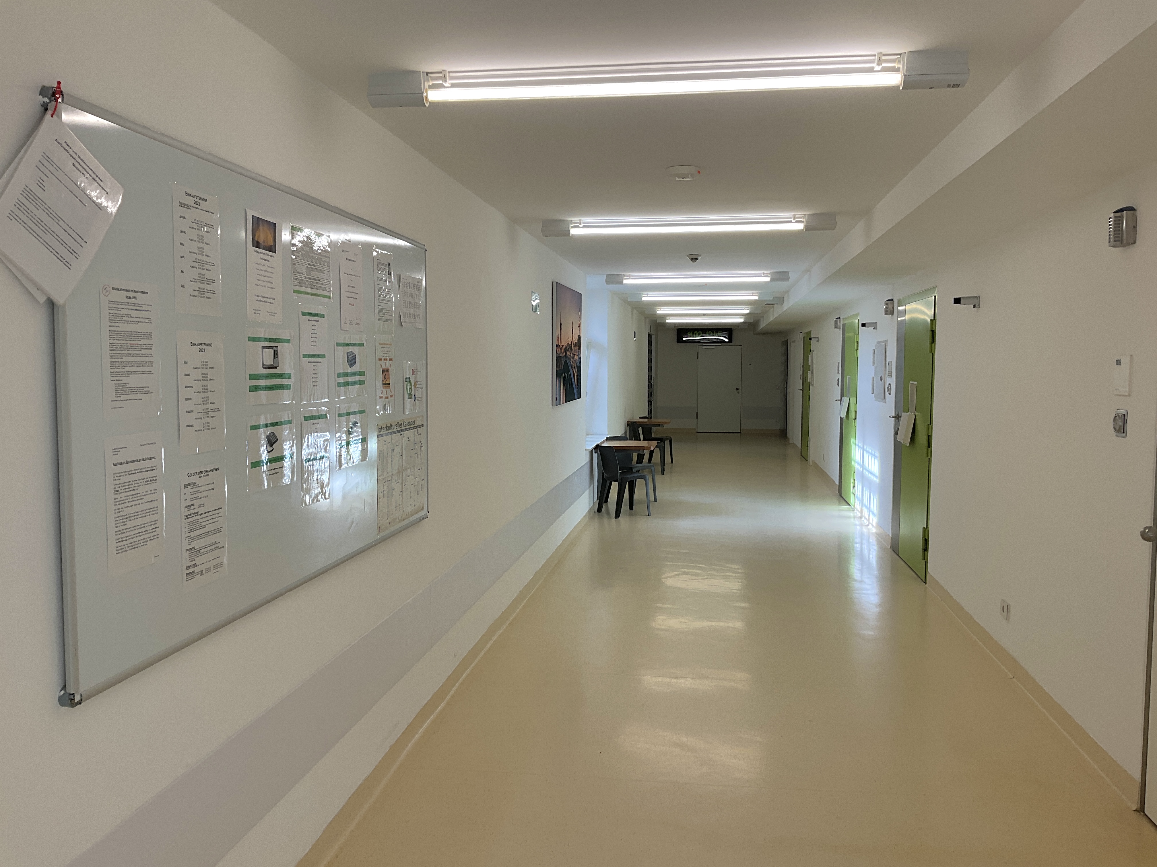 Langer Gang mit grünen Zellentüren auf der linken Seite. Tische mit Stühlen an der rechten Wand.