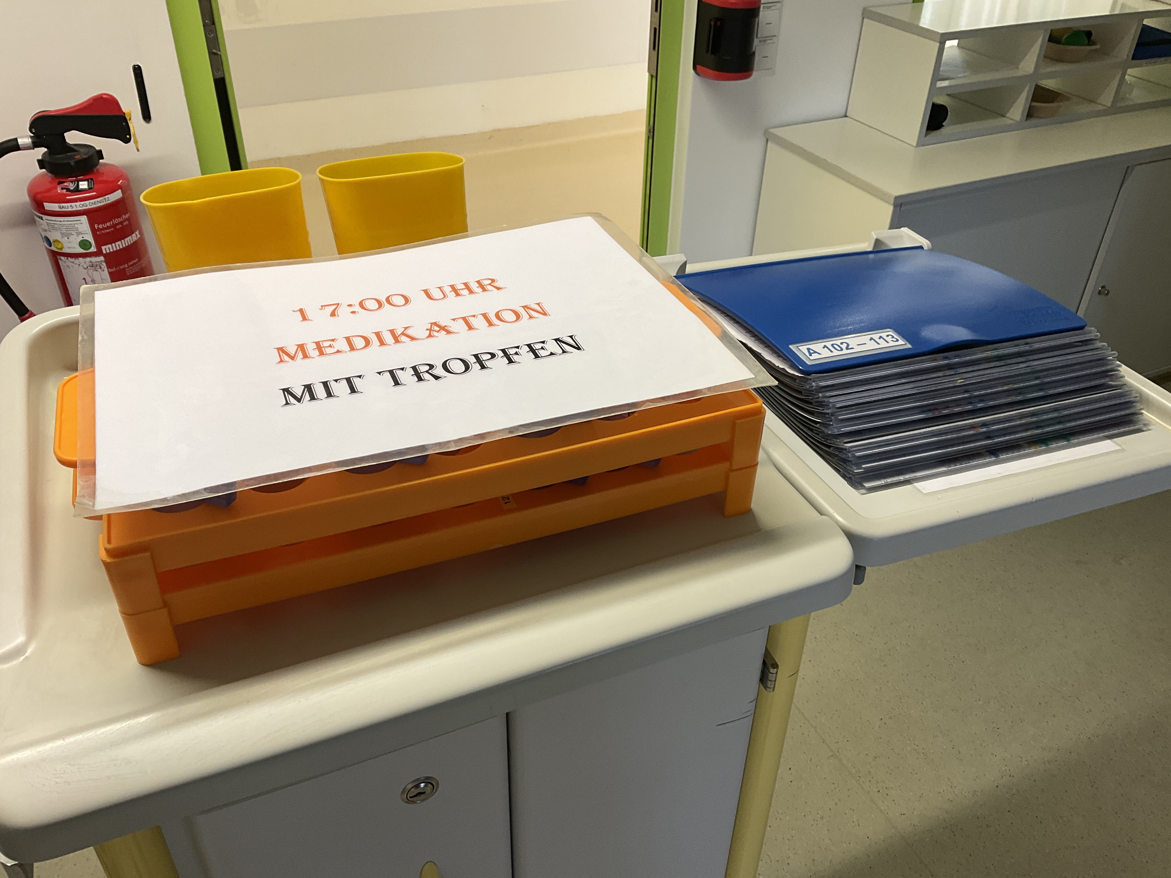 Medikamentenwagen mit orangenem Medikamententablett, daneben eine blaue Dokumentationsmappe in Stationszimmer