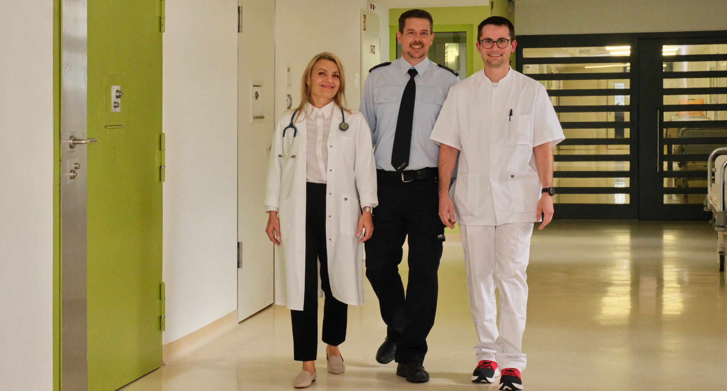 Eine Ärztin, ein Pfleger und ein Vollzugsbediensteter laufen durch eine Krankenstation.